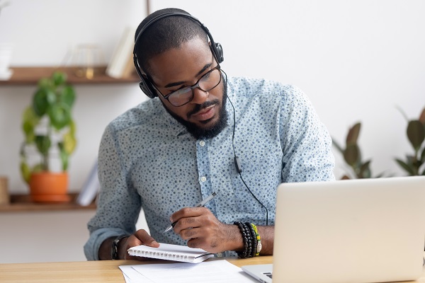 Young man at a laptop with headphone on and making notes